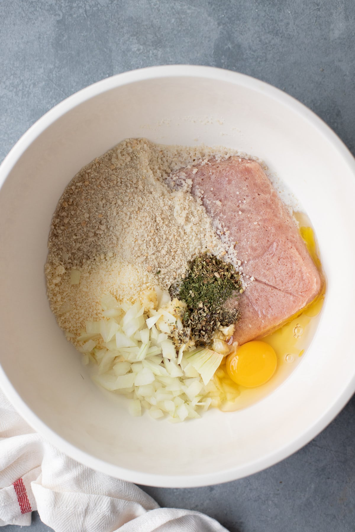 Overhead view of ground turkey, breadcrumbs, a beaten egg, grated parmesan cheese, and Italian seasoning in a large white mixing bowl.