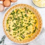 an overhead shot of bacon quiche with fresh herbs on top with eggs and cheese on the side on a white background