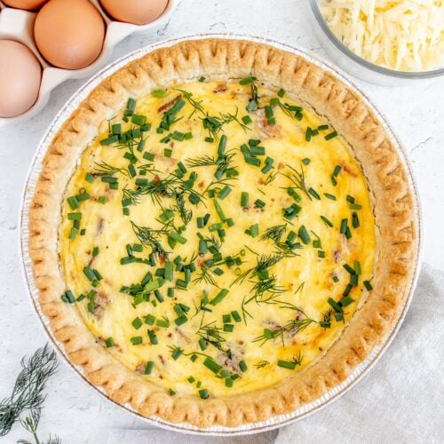 an overhead shot of bacon quiche with fresh herbs on top with eggs and cheese on the side on a white background