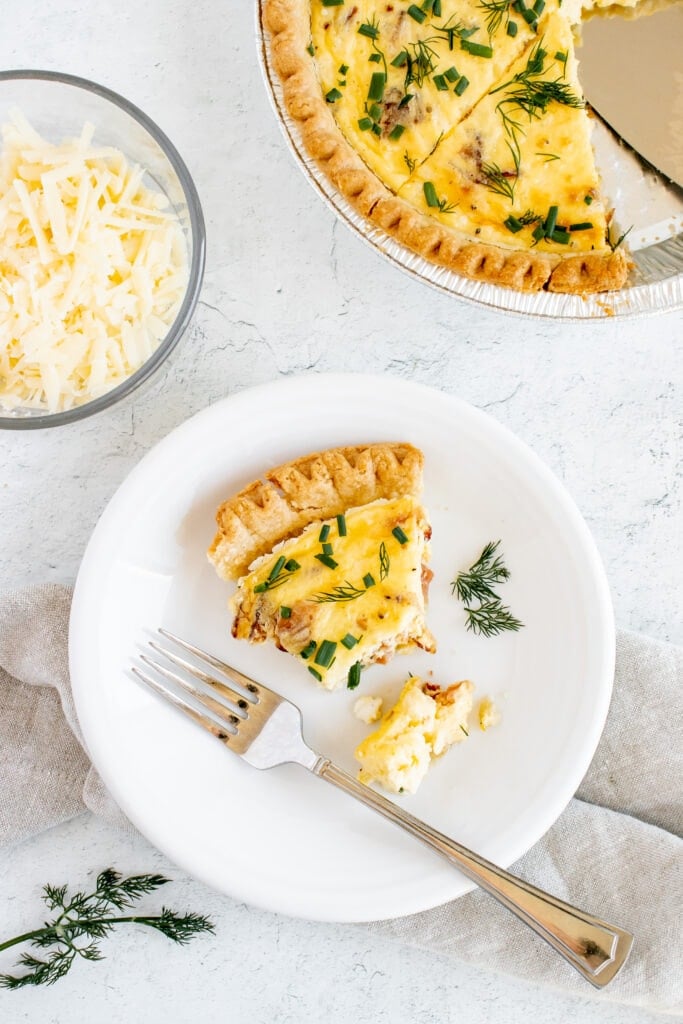 an overhead shot of a piece of bacon cheese quiche on a white plate with a bite taken out of it with a pan of quiche on the side