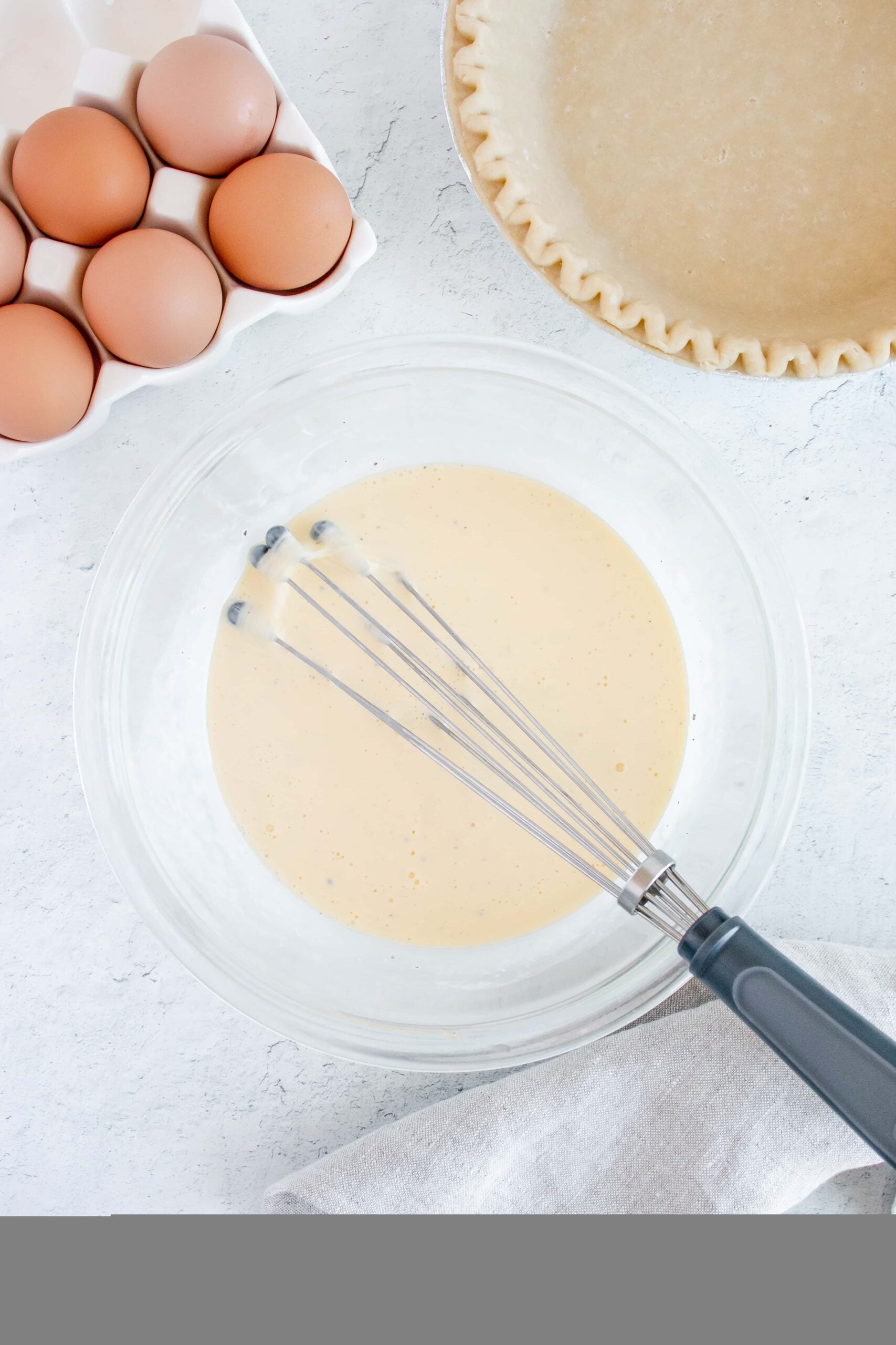 egg mixture (eggs, milk, and spices) beaten in a clear medium bowl for bacon quiche with a pie crust and eggs on the side