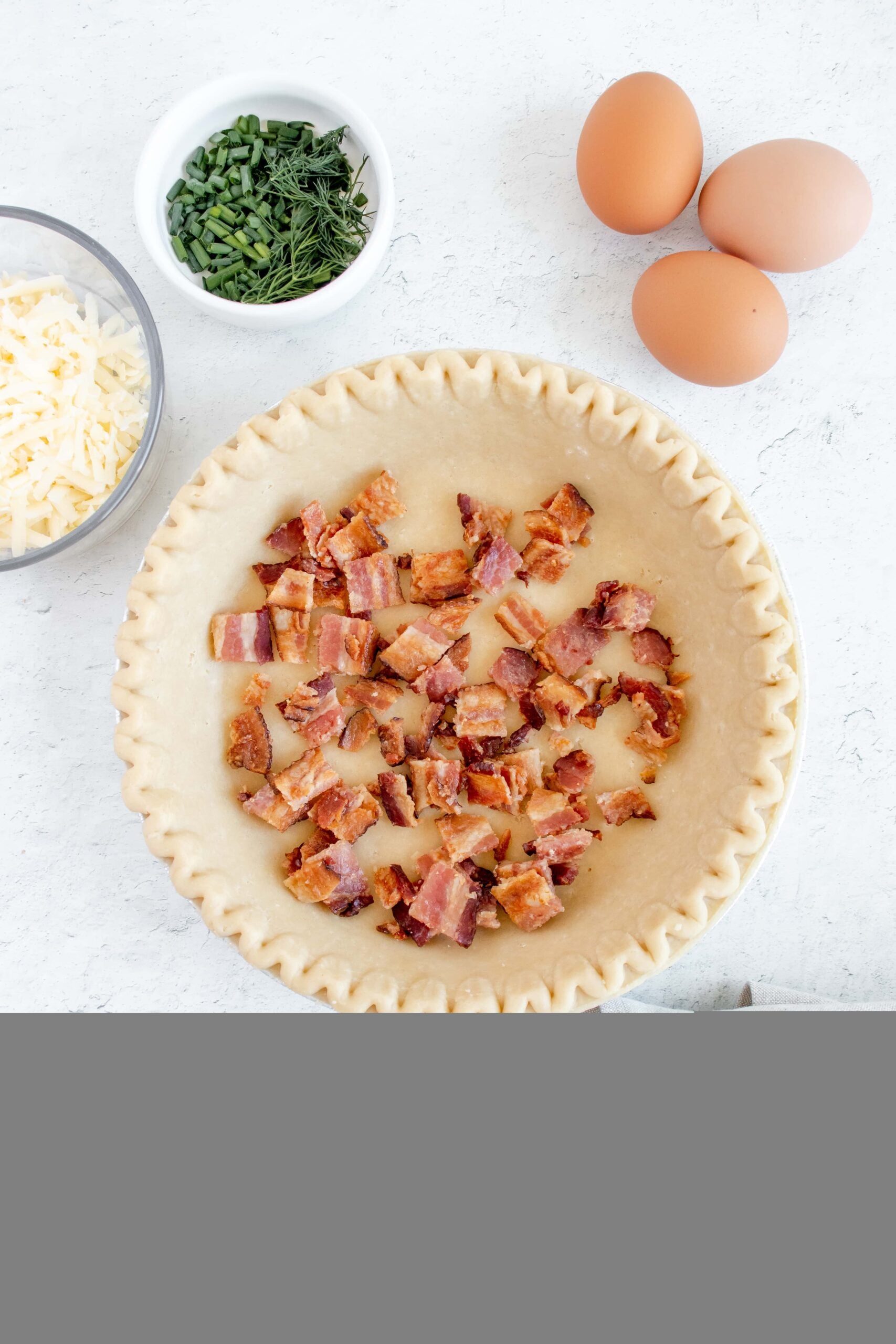 bacon crumbled in a pie crust for bacon and cheese quiche with eggs, cheese, and fresh herbs on the side