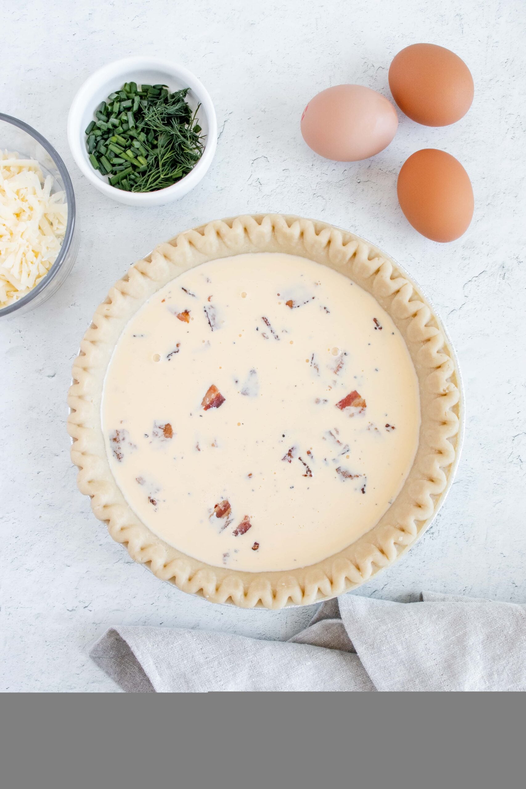 ingredients for bacon and cheese quiche uncooked in a pie crust with eggs, cheese, and fresh herbs on the side
