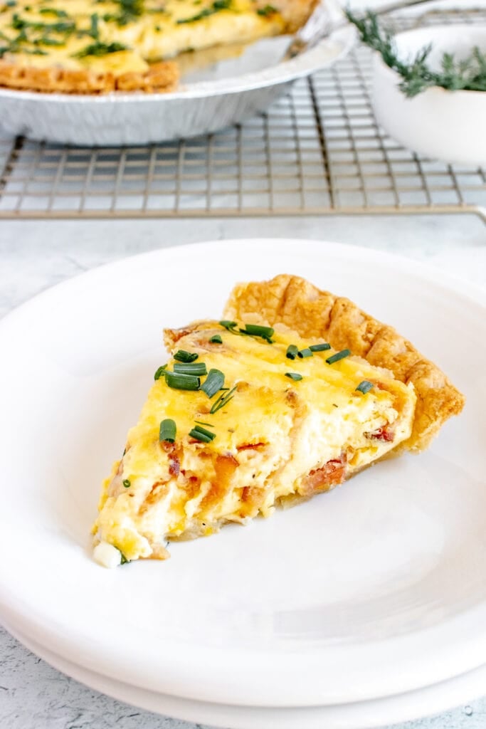 a piece of bacon quiche on a white plate with a pan of quiche in the background
