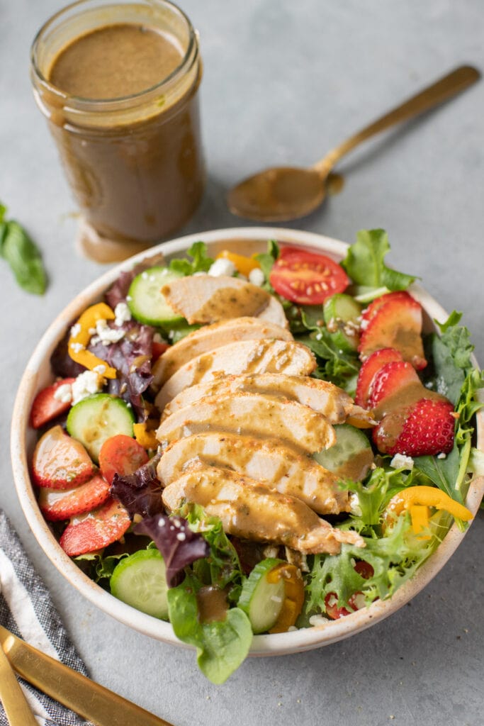 creamy balsamic dressing on a salad with sliced grilled chicken, cucumbers, tomatoes and strawberries on top with a jar of dressing in the background