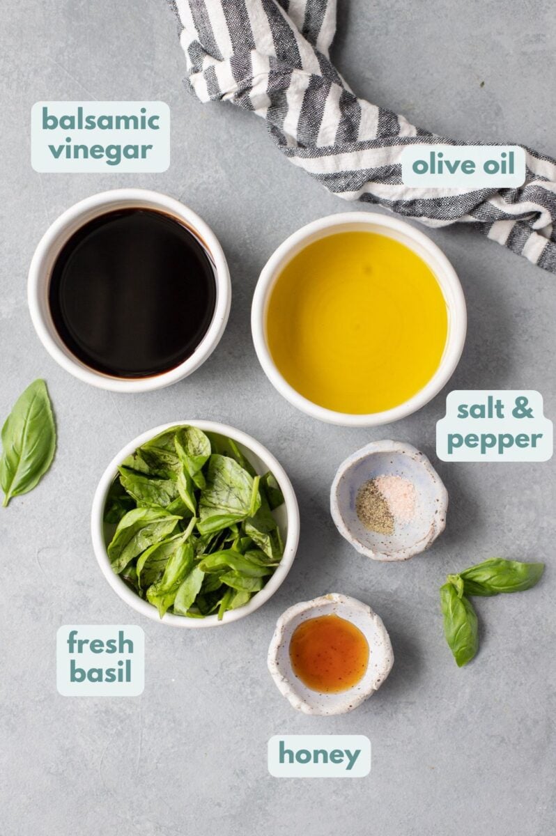 Ingredients for creamy balsamic dressing in white bowls on a grey background: olive oil, balsamic dressing, fresh basil, honey, salt & pepper