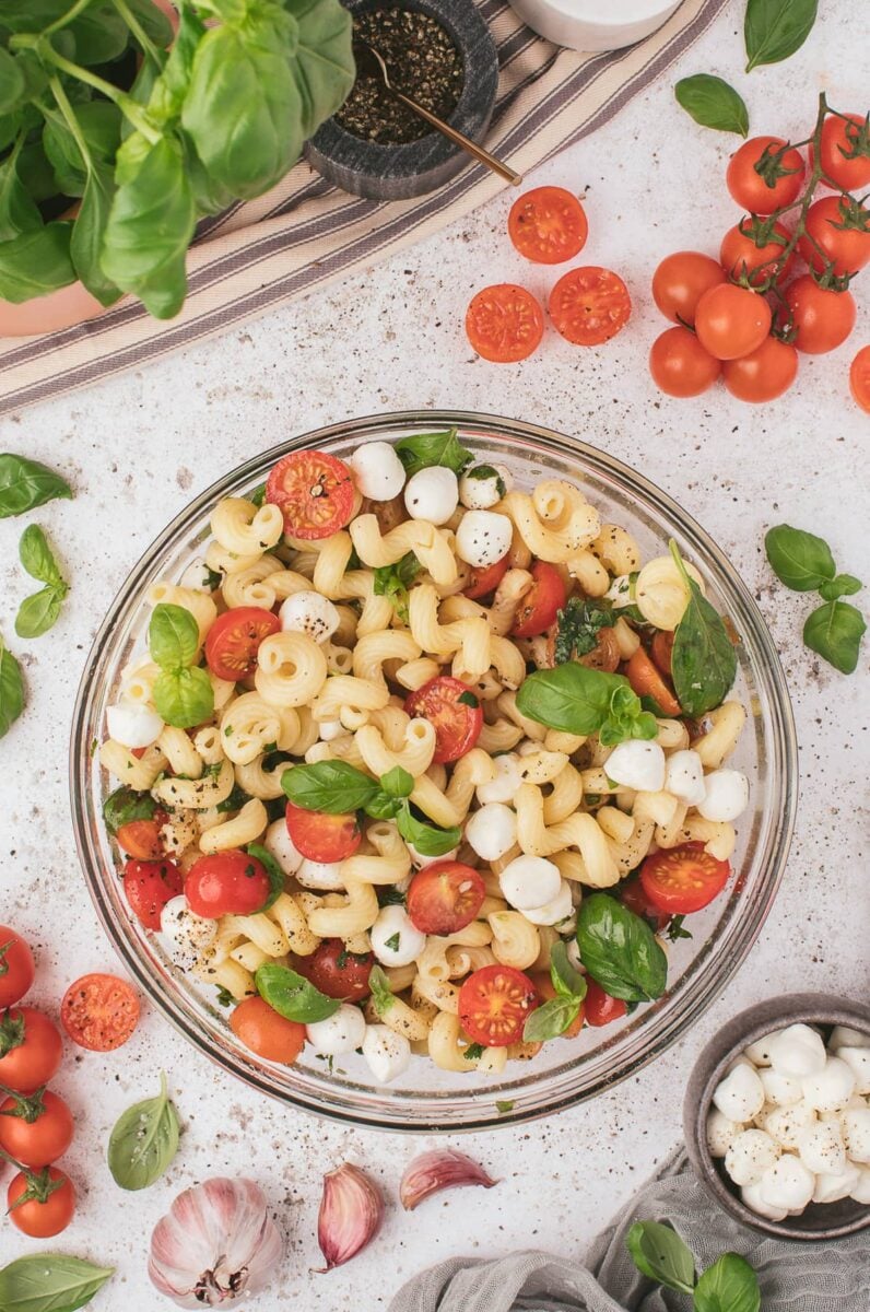 A large bowl of caprese pasta salad with mozzarella pearls and tomatoes on the side