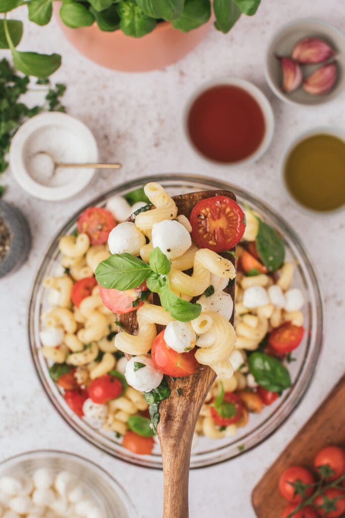 a close up of a wooden spoon with caprese pasta salad and a bowl of the pasta salad in the background