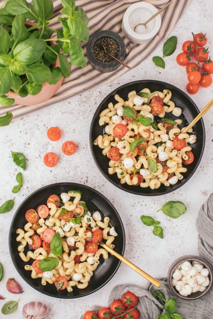 An overhead shot of two black plates of caprese pasta salad with tomatoes, basil and mozzarella pearls on the side