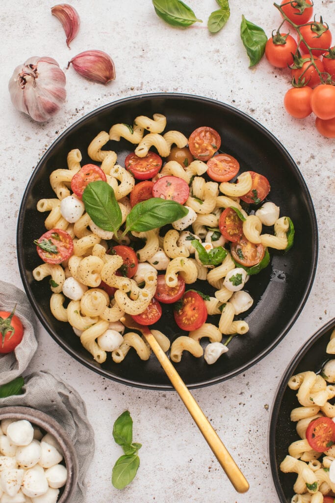 A black plate of caprese pasta salad with a gold fork on a grey background