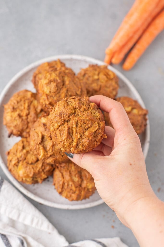A hand holding a healthy carrot muffin over a plate of carrot cake muffins
