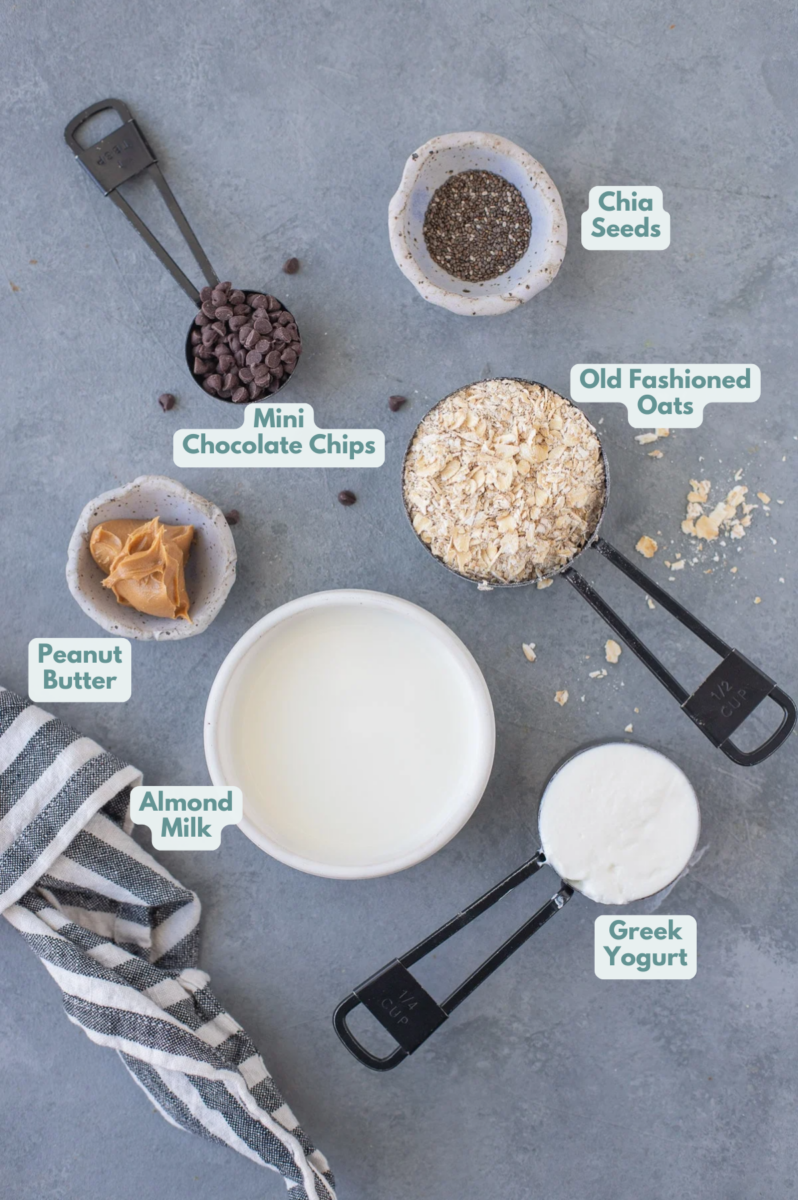 Ingredients for peanut butter overnight oats in white bowls on a grey background: peanut butter, chia seeds, almond milk, chocolate chips