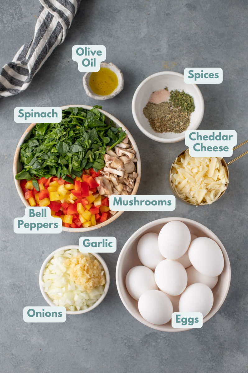 Ingredients for healthy breakfast casserole in white bowls: olive oil, garlic, onions, bell peppers, mushrooms, chopped spinach, eggs, dried parsley, dried basil, dried oregano, pepper, salt, cheddar cheese