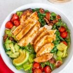An overhead shot of a white bowl of Honey Mustard Chicken Salad with tomatoes, avocado and bacon on a bed of mixed greens