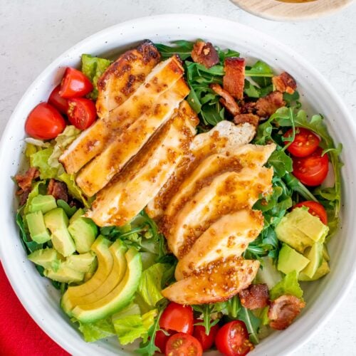 An overhead shot of a white bowl of Honey Mustard Chicken Salad with tomatoes, avocado and bacon on a bed of mixed greens