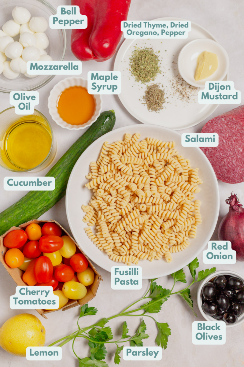 Ingredients for Italian pasta salad in white bowls, viewed from overhead: fusilli pasta, mini mozzarella balls, cherry tomatoes, red onion, cucumber, black olives, red bell pepper, fresh parsley, salami, dried thyme, dried oregano, black pepper, lemon juice, olive oil, Dijon mustard, and maple syrup