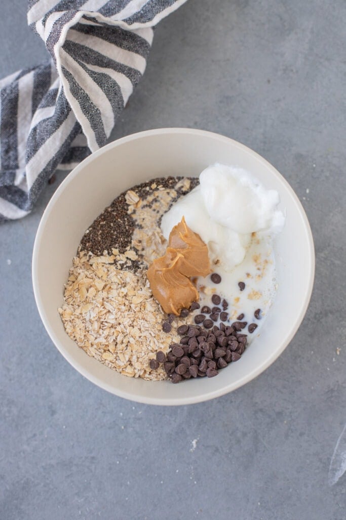 All ingredients for peanut butter overnight oats in a big white bowl on a grey background