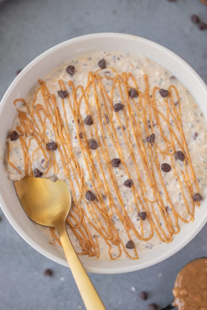 A close up of peanut butter overnight oats with chocolate chips and peanut butter drizzle on top with a gold spoon in it