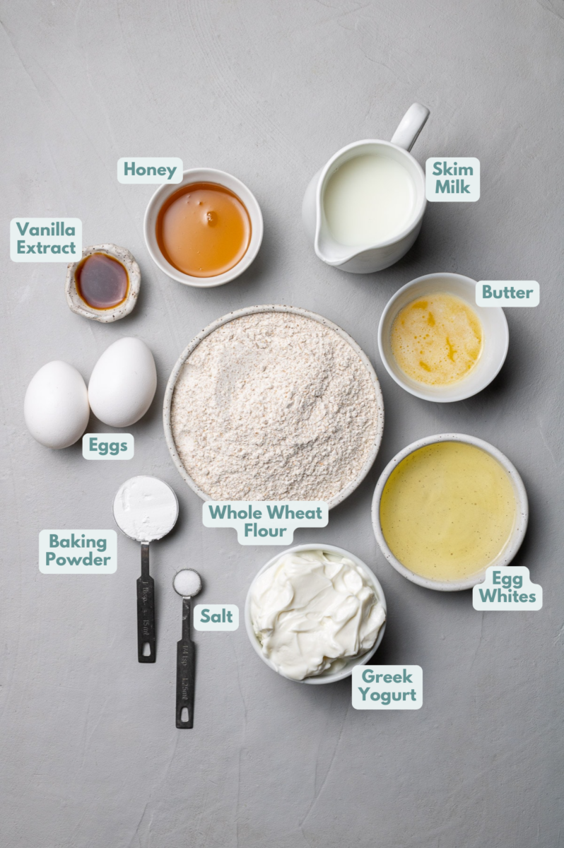 Ingredients for protein waffles in white bowls: nonfat Greek yogurt, skim milk, eggs, egg whites, honey, vanilla extract, butter, whole wheat flour, baking powder, salt
