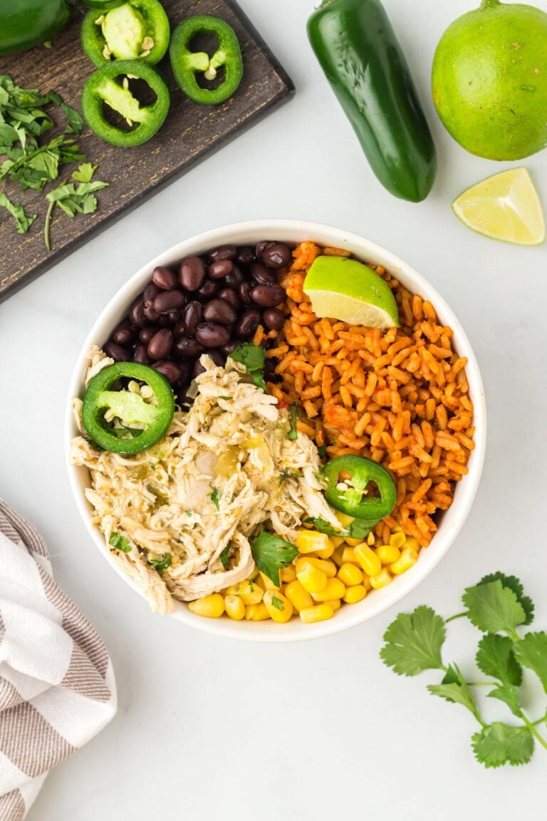 an overhead shot of salsa verde chicken in a white bowl with black beans, corn, and rice with cilantro, jalapeños and limes on the side