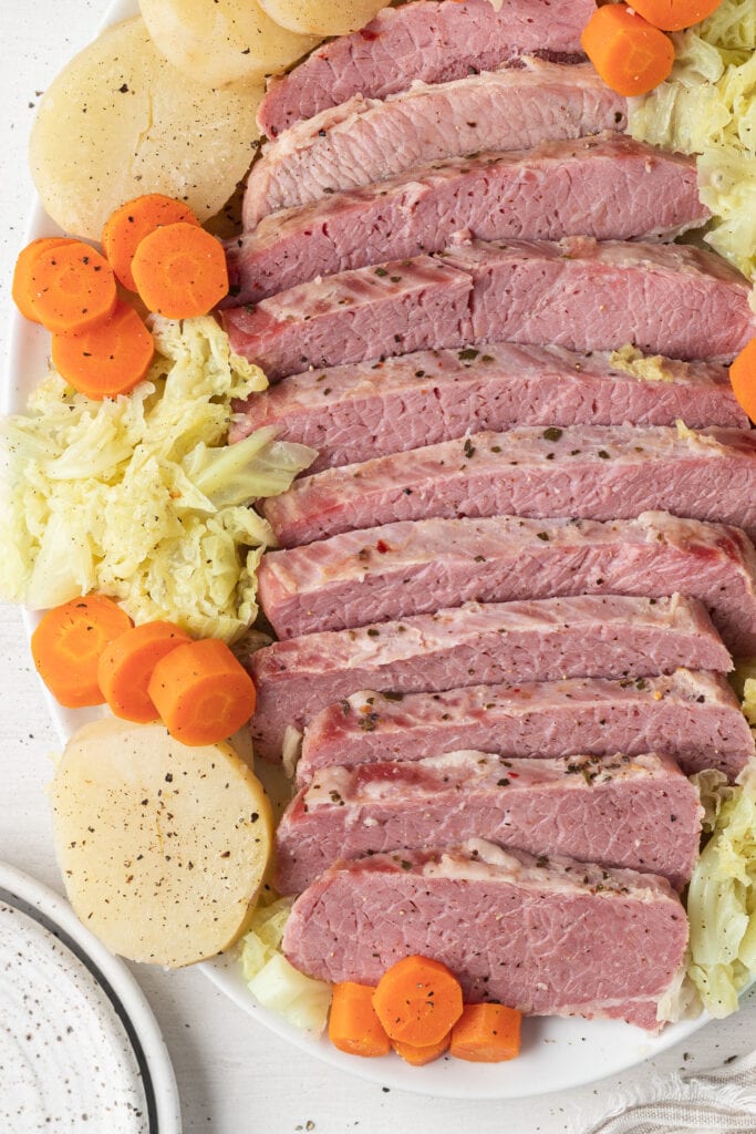 a close up of slow cooker corned beef on a white platter surrounded by cabbage, carrots and potatoes