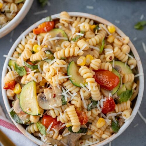 an overhead shot of pasta primavera with parmesan cheese on top in a white bowl on a grey background with a pan and bowl of pasta on the side