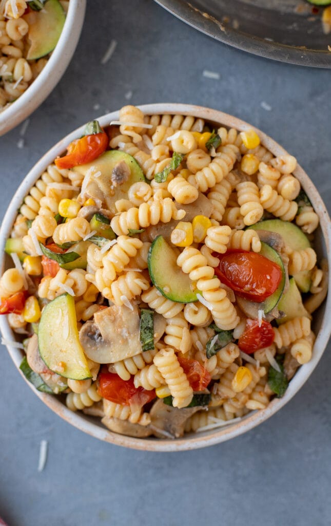 an overhead shot of pasta primavera in a white bowl on a grey background with a pan and bowl of pasta on the side