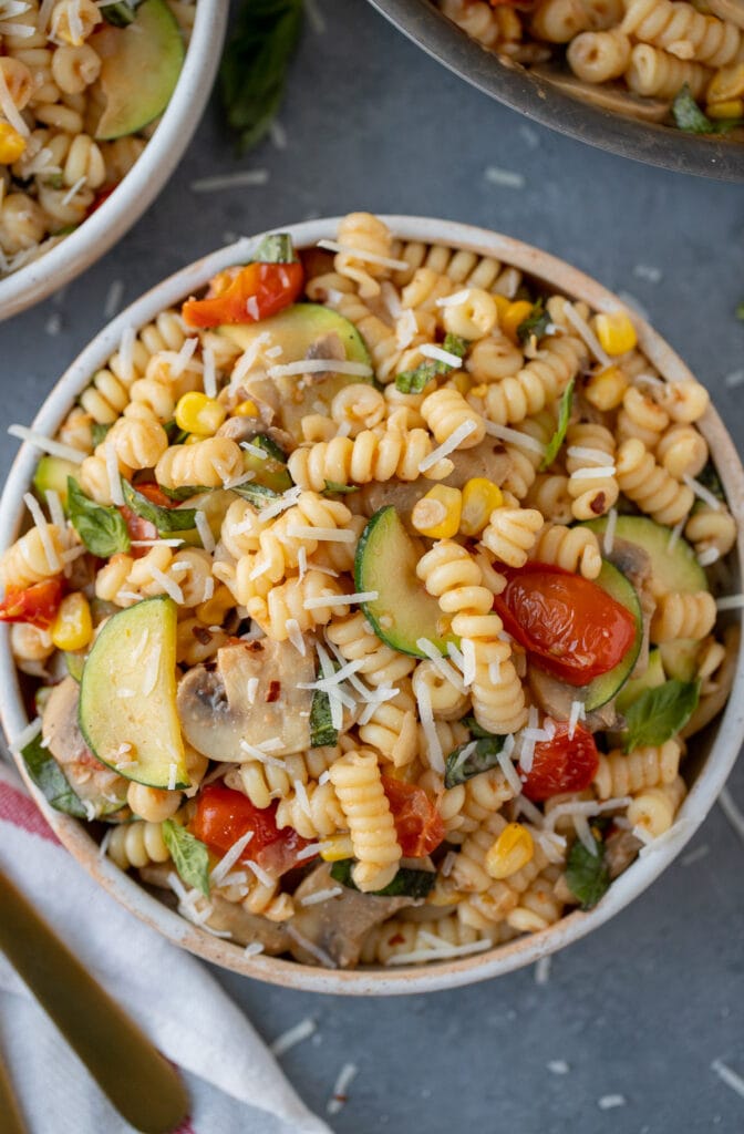 a white bowl of pasta primavera with parmesan cheese on top on a grey background with a pan and bowl of pasta on the side