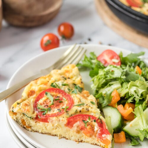 A slice of vegetable frittata on a white plate with a salad on the side