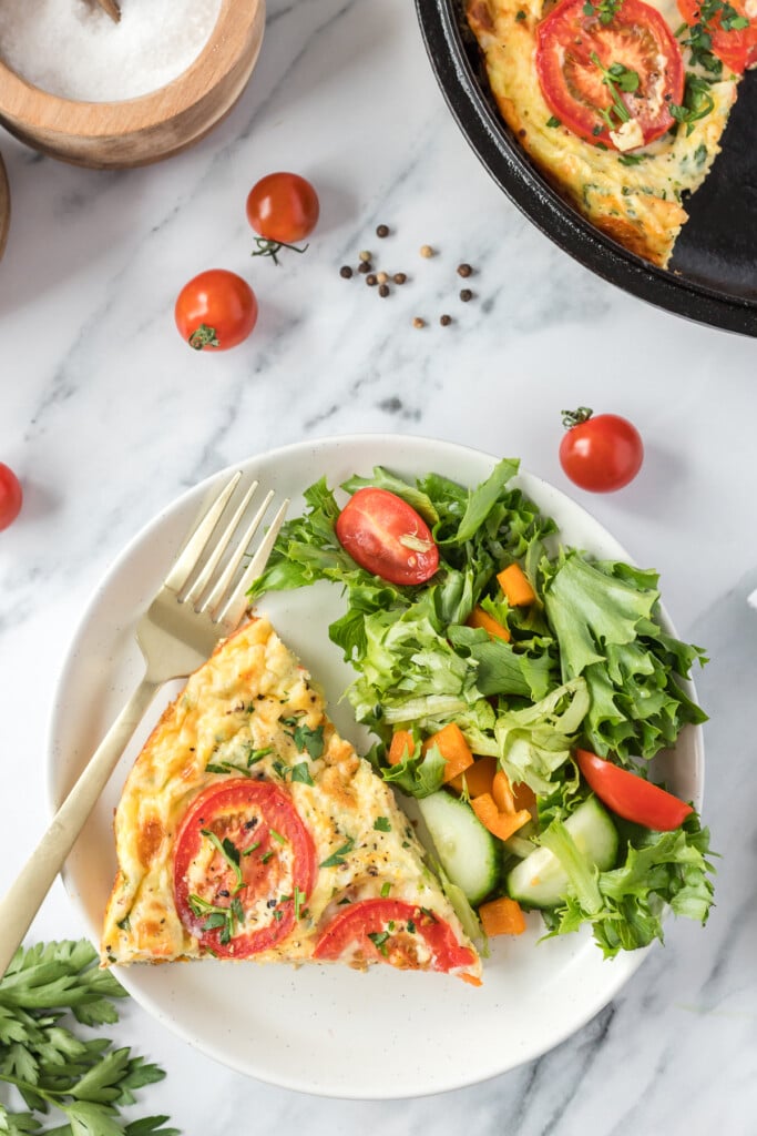 An overhead shot of a white plate with a piece of vegetable frittata and a salad on it with a cast iron pan of frittata on the side