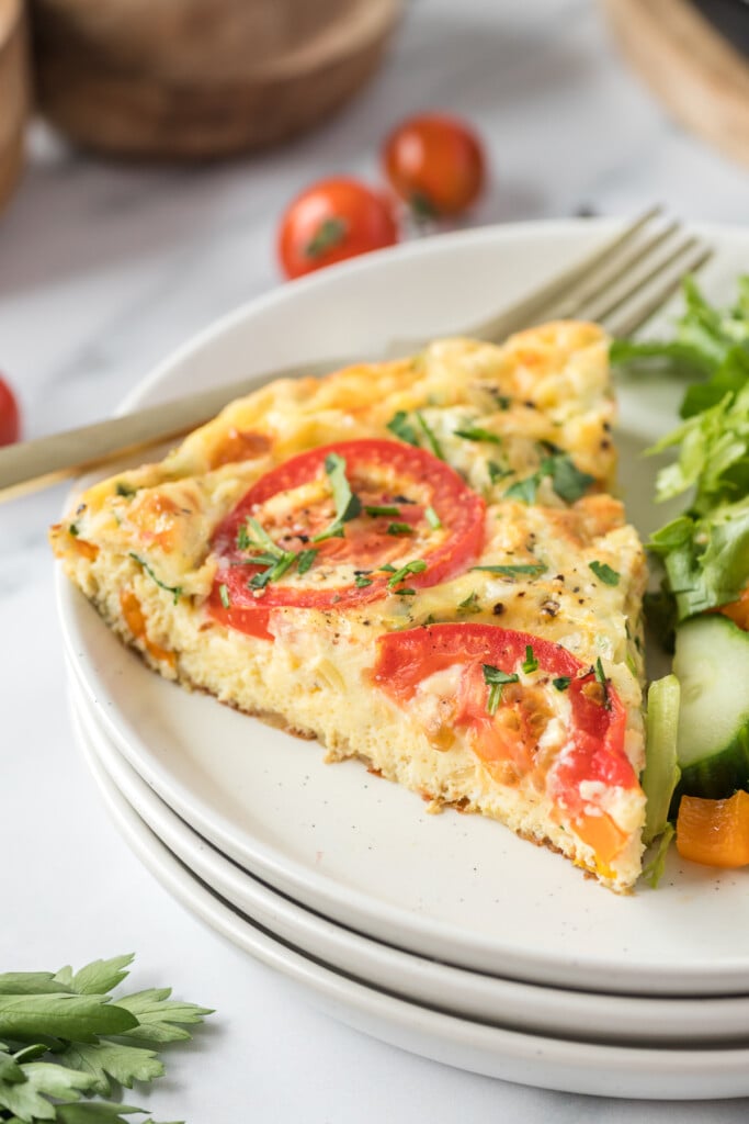 A piece of vegetable frittata on a white plate with a fork and salad in the background