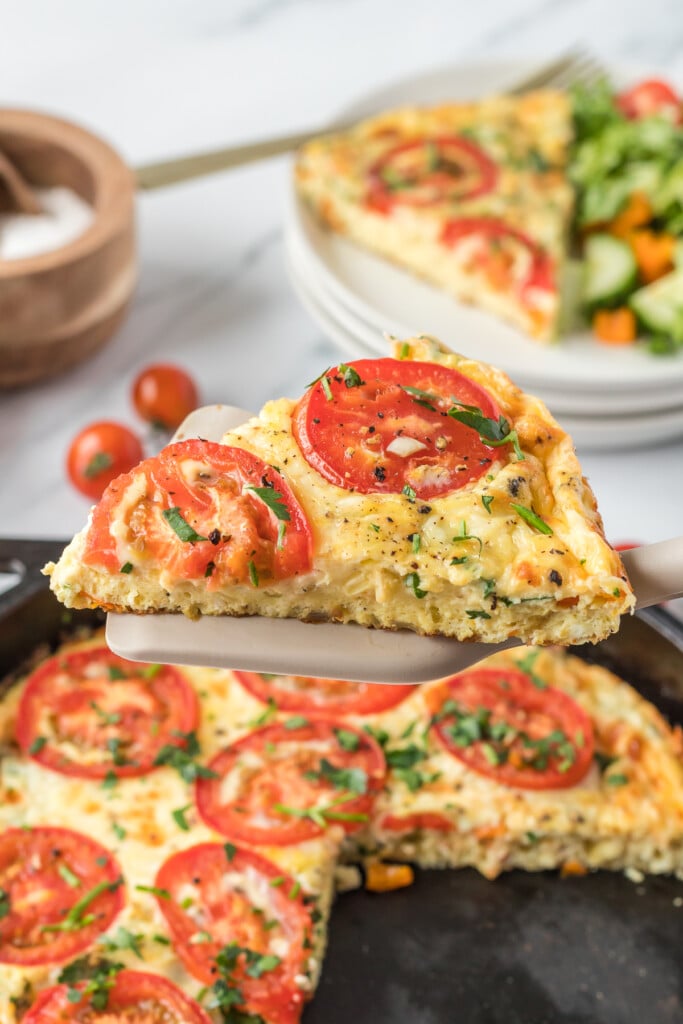 a piece of vegetable frittata being lifted out of a pan of the frittata with a plate in the background