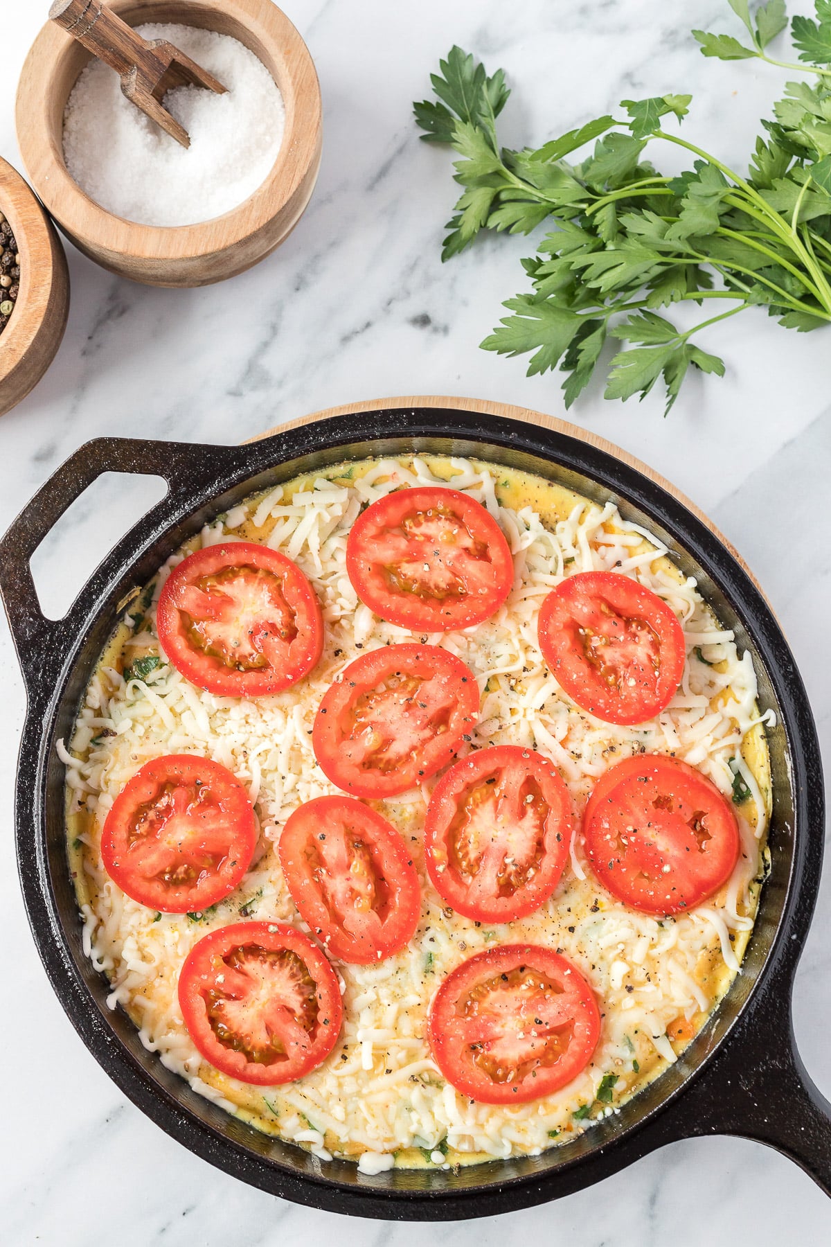 vegetable frittata in a cast iron skillet with uncooked/melted cheese and tomatoes on top