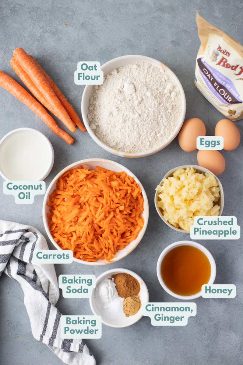Ingredients to make healthy carrot muffins in white bowls: shredded carrots, crushed pineapple, eggs, honey, coconut oil, gluten free oat flour, baking powder, baking soda, salt, cinnamon, and ground ginger.