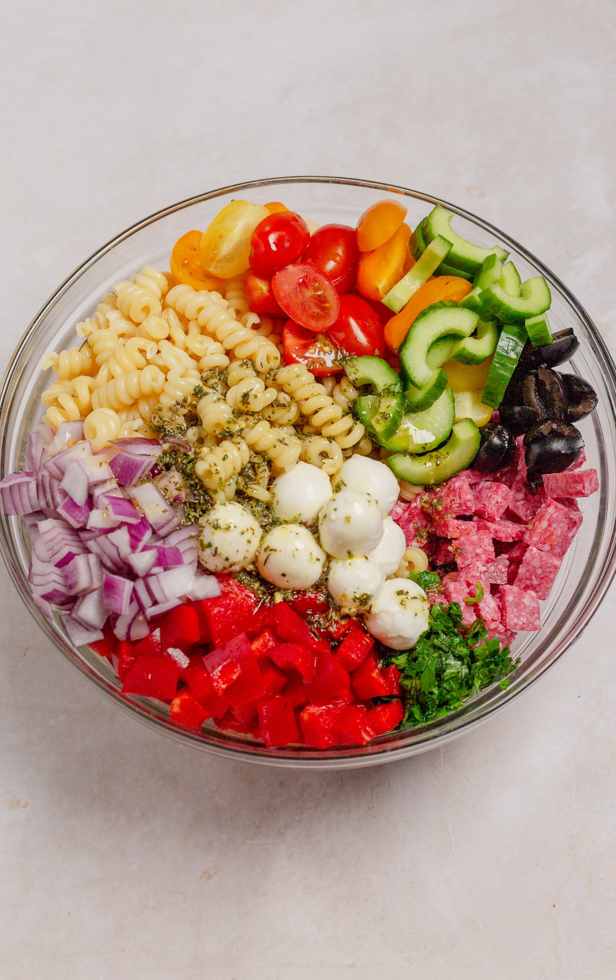 Chopped and sliced vegetables with  diced salami, black olives, and mozzarella cheese balls in a large glass mixing bowl, viewed from overhead.