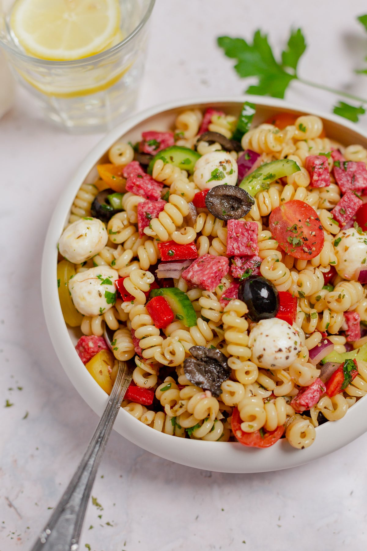 Large white bowl full of Italian pasta salad with salami, mozzarella balls, red bell pepper, cherry tomatoes, and black olives.