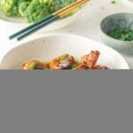 A white bowl of Honey Garlic Shrimp with scallions on top and a bowl of broccoli in the background