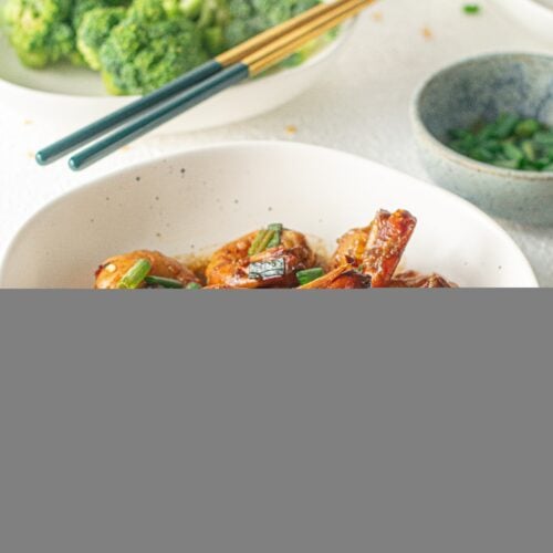 A white bowl of Honey Garlic Shrimp with scallions on top and a bowl of broccoli in the background