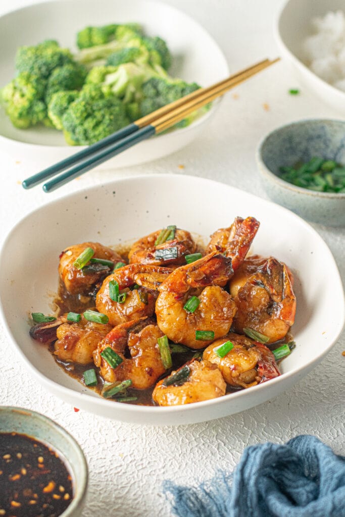 A white bowl of Honey Garlic Shrimp with scallions on top and a bowl of broccoli in the background