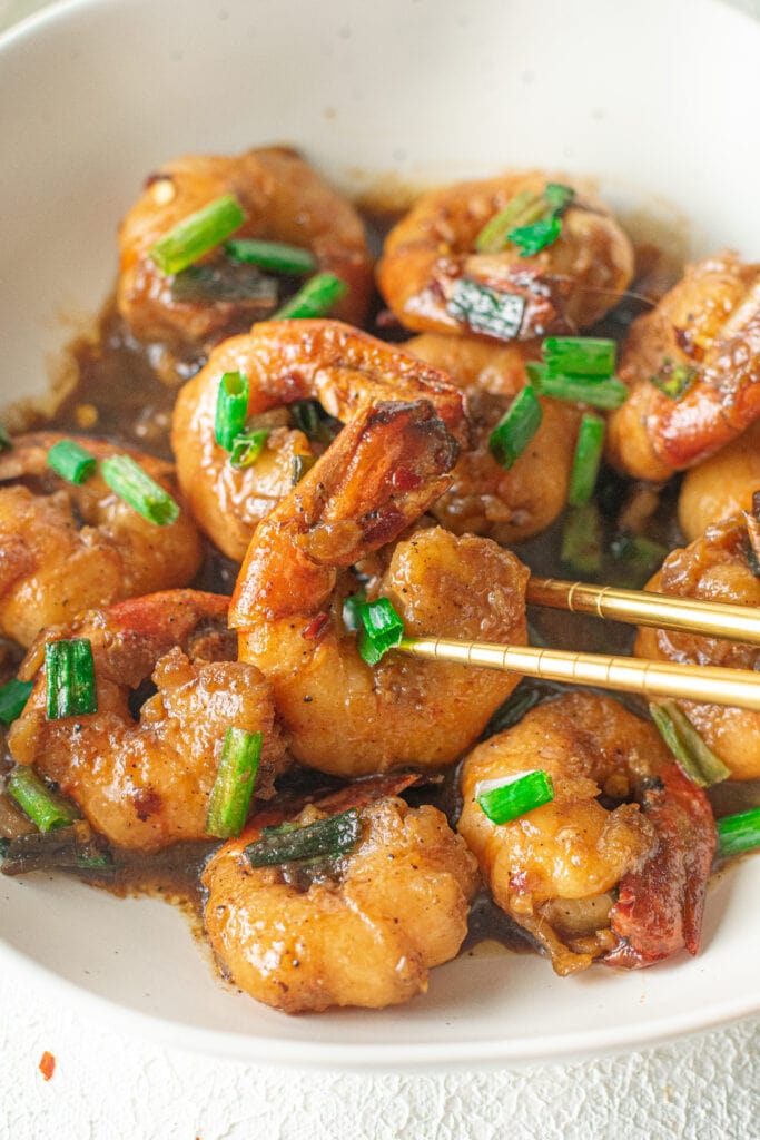 A close up of a white bowl of honey garlic shrimp with green scallions on top with a shrimp being grabbed by gold chopsticks