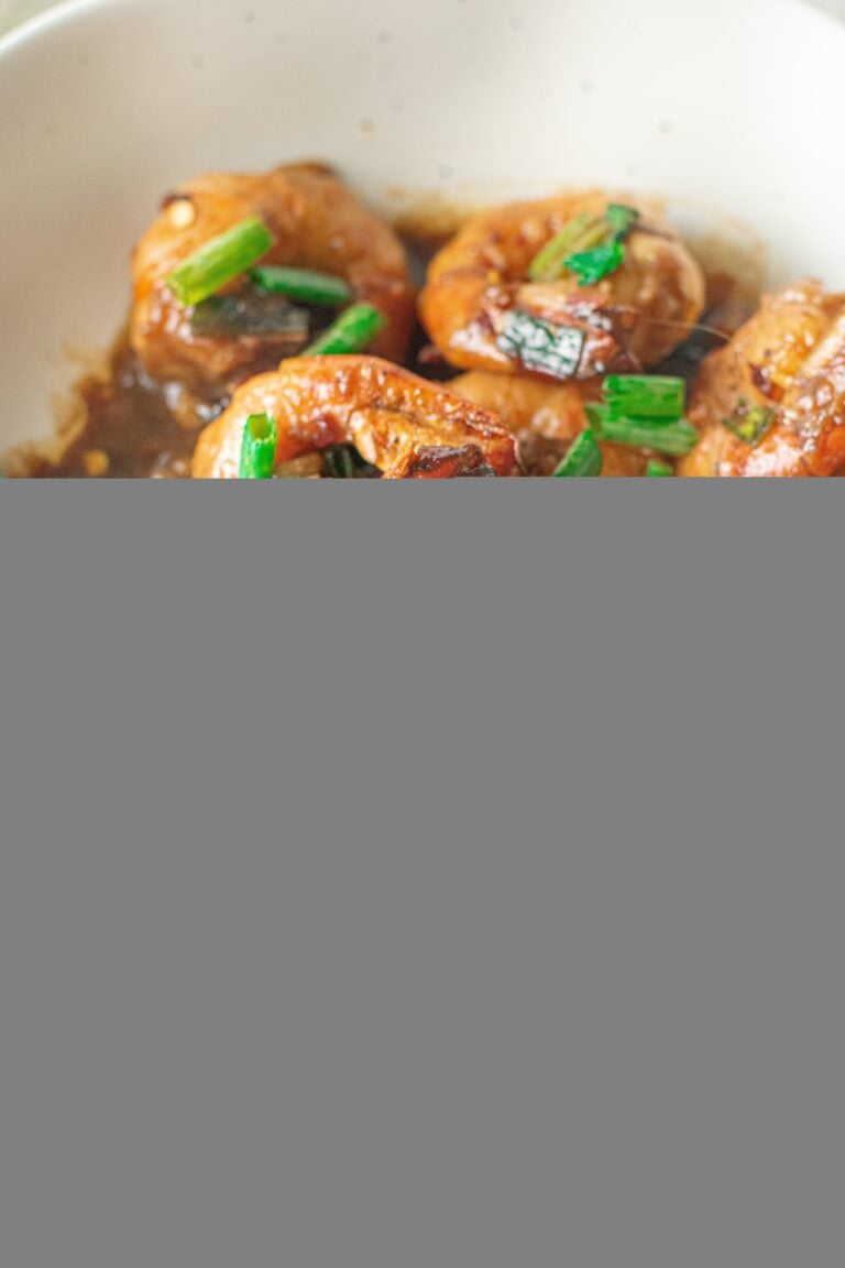 A close up of a white bowl of honey garlic shrimp with green scallions on top with a shrimp being grabbed by gold chopsticks