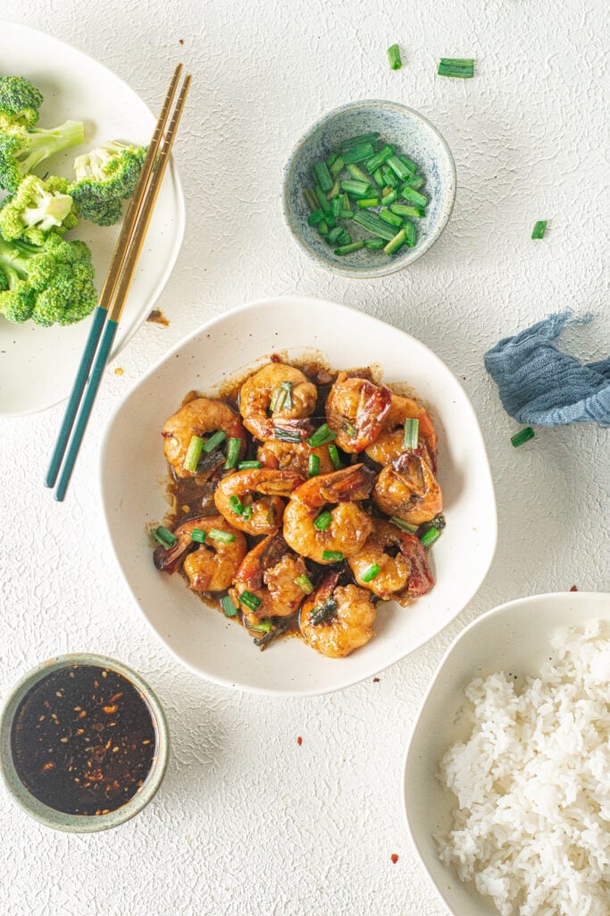 An overhead shot of honey garlic shrimp in a white bowl with scallions, broccoli, and rice on the side