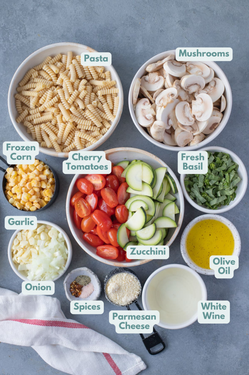 Ingredients for pasta primavera in white bowls on a grey background: olive oil, onion, garlic, red pepper flakes, salt, pepper, cherry tomatoes, mushrooms, zucchini, frozen corn, Parmesan cheese, white wine, pasta, fresh basil