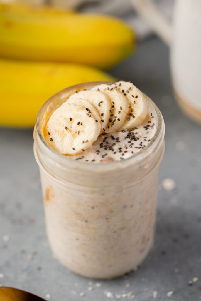 A mason jar with banana oats with banana slices and chia seeds on top and bananas in the background