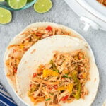 An overhead shot of two tortilla with crock pot chicken fajitas on a grey background with limes and a slow cooker on the side