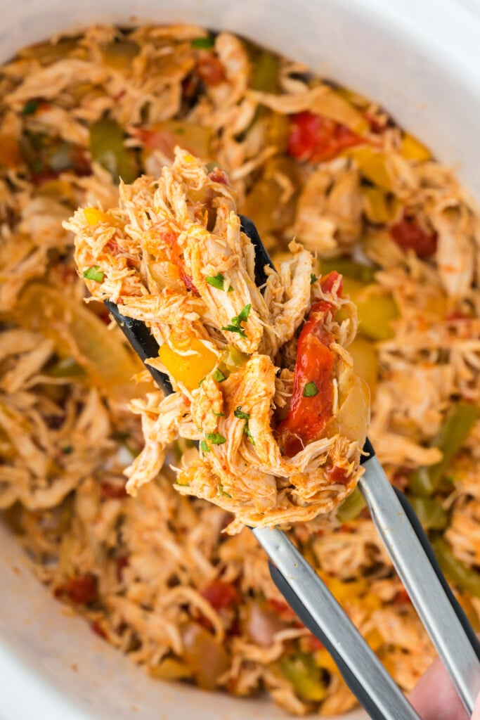 Tongs taking a scoop of crock pot chicken fajitas out of the slow cooker in the background