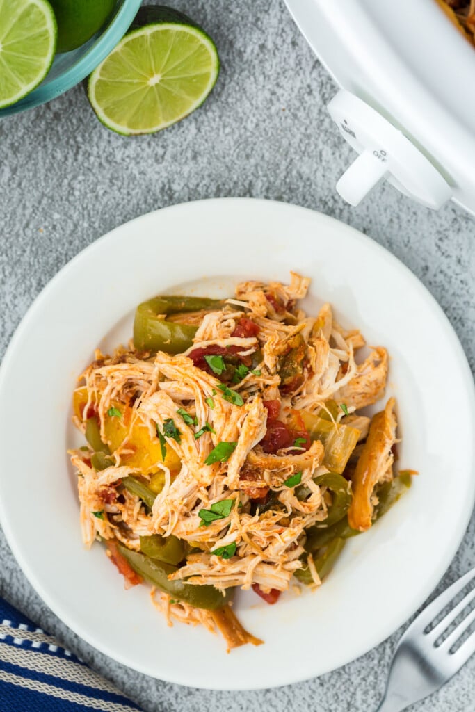 An overhead shot of a white plate of crockpot chicken fajitas with a slow cooker and limes on the side