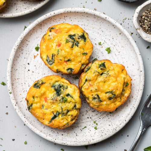 An overhead shot of a plate of three egg muffin cups with more on the side on a grey background