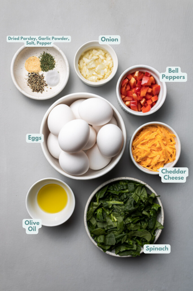 Ingredients for healthy egg muffin cups in white bowls on a grey background: olive oil, spinach, bell peppers, onion, eggs, garlic powder, dried parsley, salt + pepper, cheddar cheese (optional)
