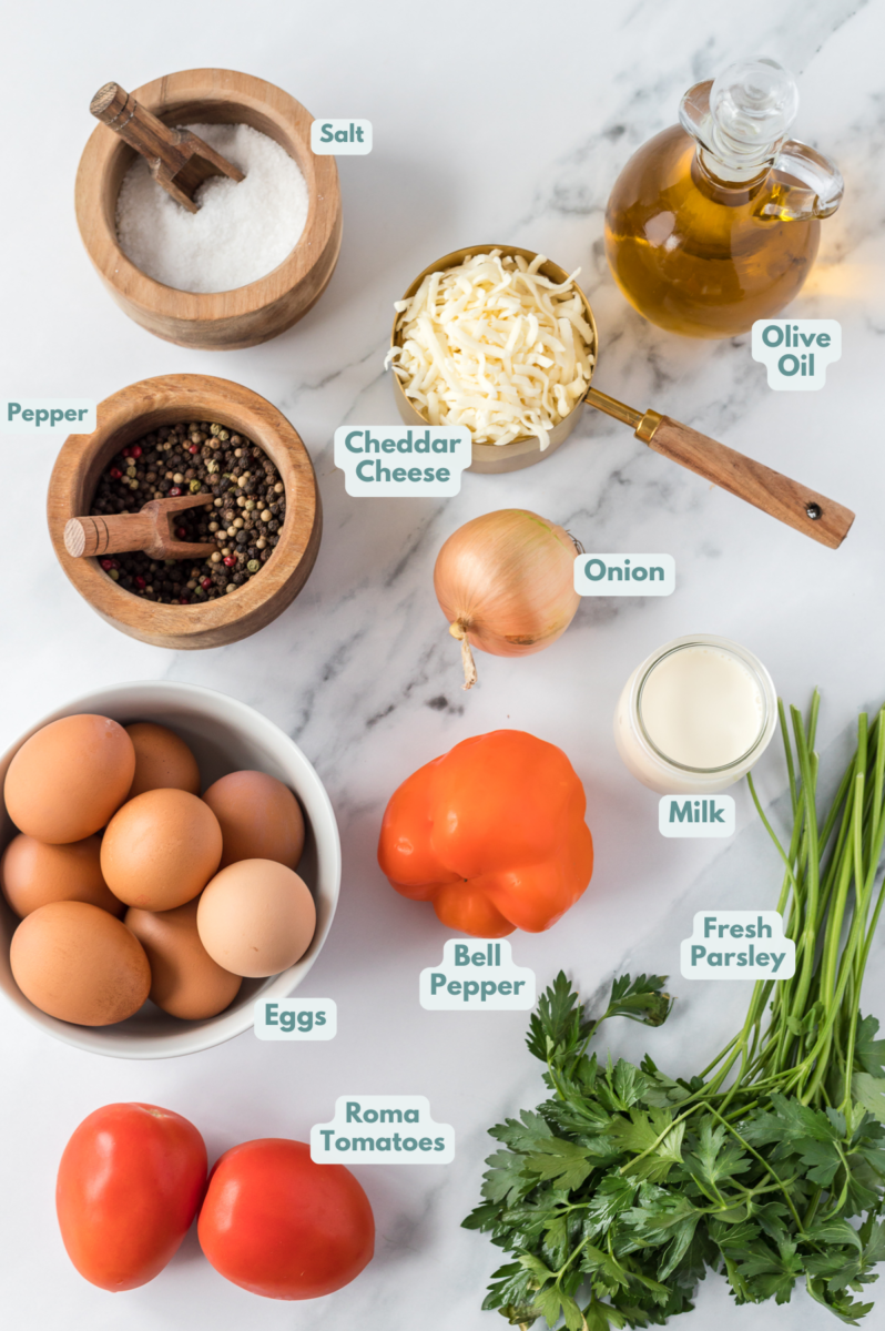 Ingredients for vegetable frittata laid out on a marble background: eggs, milk, olive oil, bell pepper, onion, tomatoes, cheddar cheese, fresh parsley, salt and pepper
